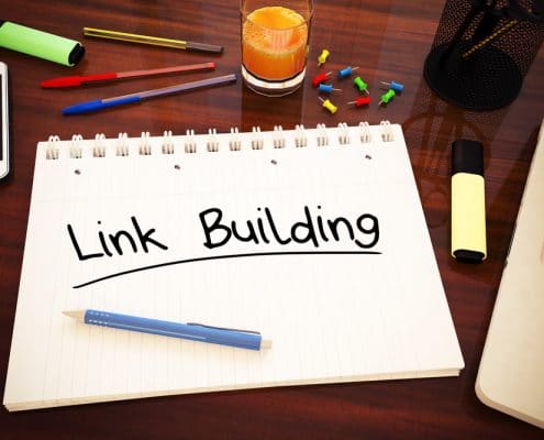 The words "Link Building" written on a notebook that is sitting on a desk with a computer and smartphone.