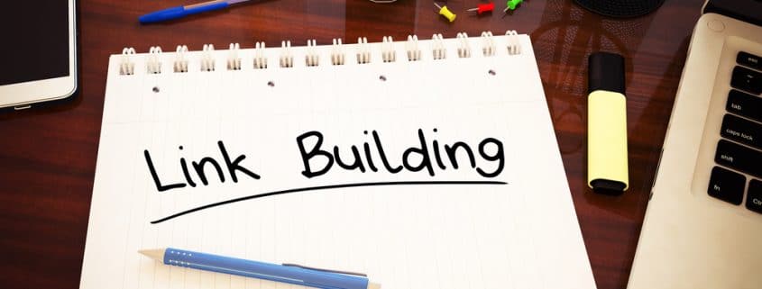 The words "Link Building" written on a notebook that is sitting on a desk with a computer and smartphone.