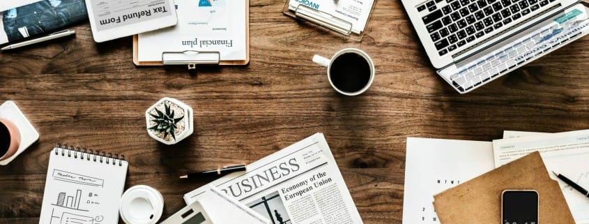 Tools for conducting business on a wooden desk including a laptop, smartphone, clipboard, newspaper, calendar, and coffee.