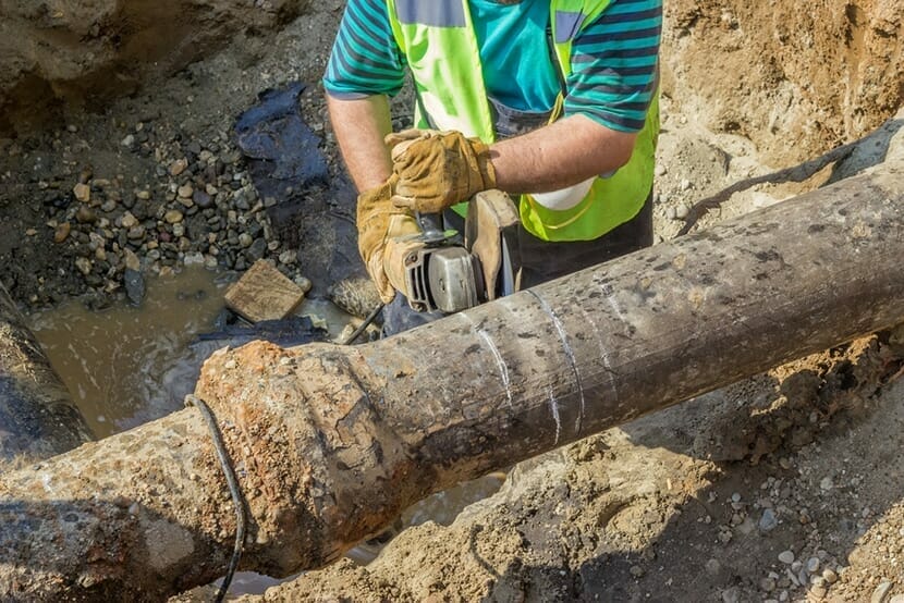 Image of a man in a ditch cutting a sewer pipe with a metal cutter.