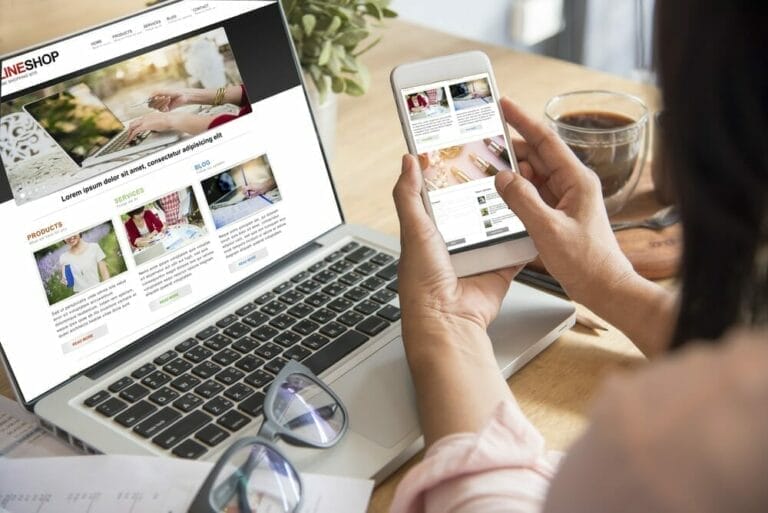Woman at a computer holding a cell phone and both are showing the same website