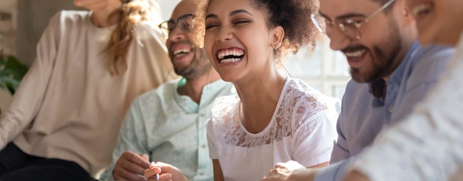 A group of people sit together laughing. The focus is on a woman in a white top smiling broadly. They appear to be enjoying a casual, light-hearted moment indoors.