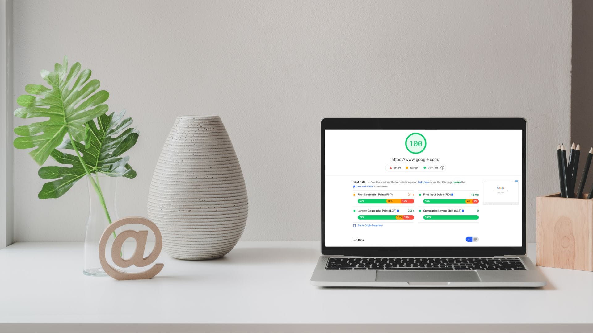 Open laptop on white desk with pencils, a couple plant leaves in a glass jar and a decorative @ symbol