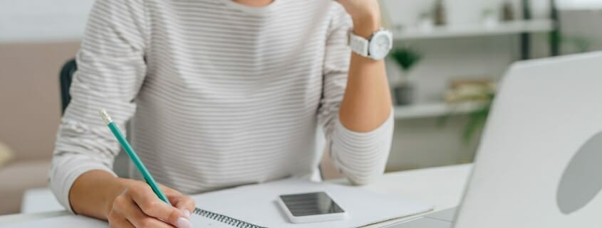 Woman with pad and pencil focused on laptop screen.