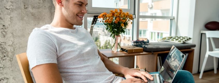 Man by a window, sitting in a chair working on his laptop