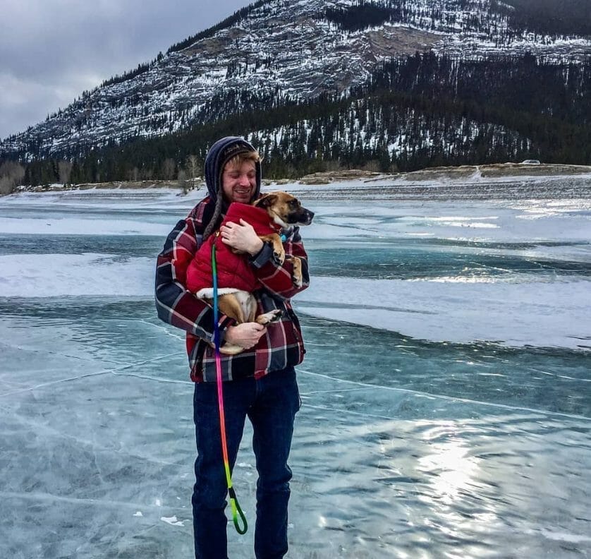 A man standing on a frozen lake holding a dog that is wearing a red dog jacket.