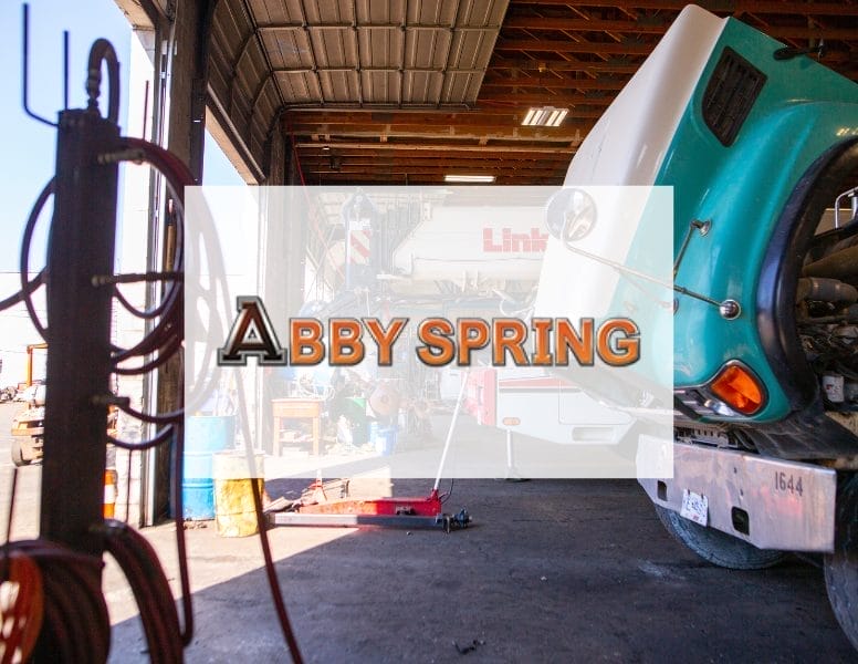 A truck repair shop with a white and green truck in the foreground, its hood open for maintenance. Tools hang on a rack nearby. Sunlight filters through the open door. Overlay text reads "Abby Spring.