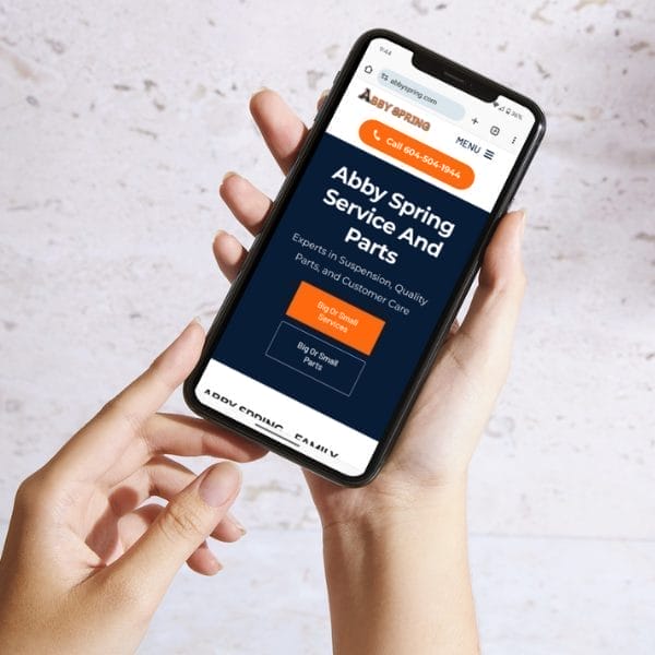 A person holds a smartphone displaying the Abby Spring Service and Parts website. The screen shows options for service information, a call button, and menu access. The phone is held with two hands over a white background.