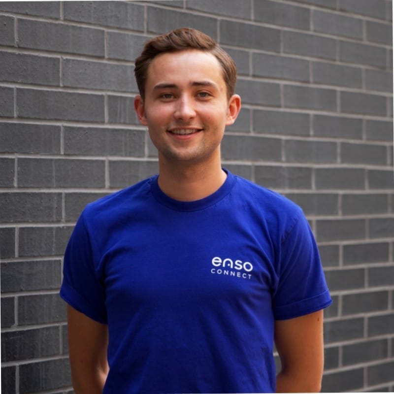 A person with short brown hair is smiling, wearing a blue T-shirt with the text "enso CONNECT" on it. They are standing in front of a gray brick wall.