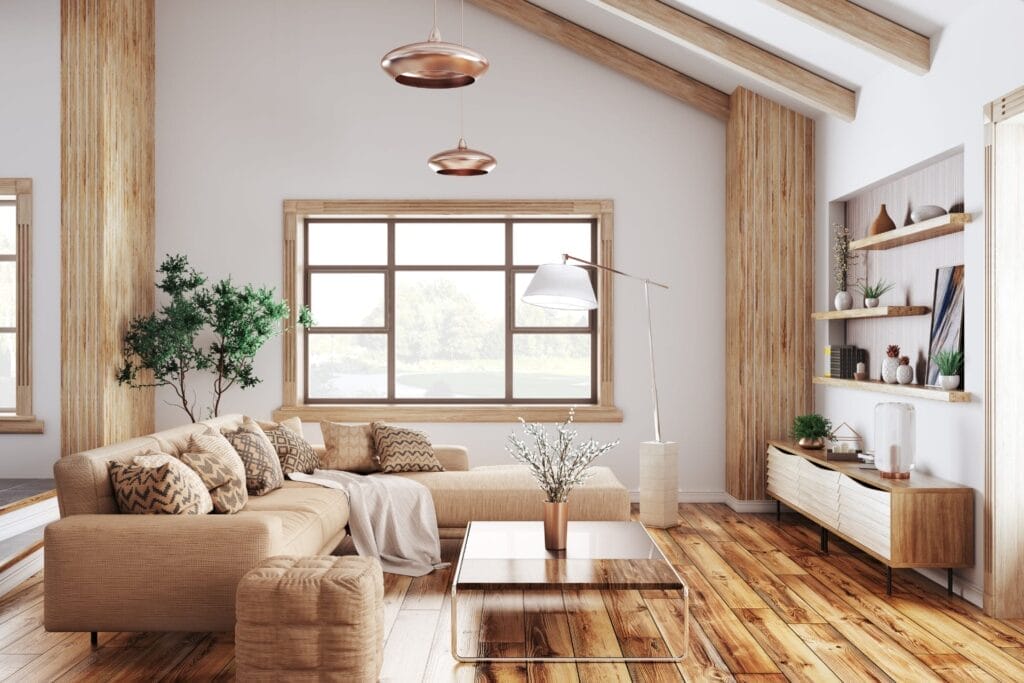 Modern living room with a beige L-shaped sofa, wooden flooring, and large windows. Features include a wooden coffee table, indoor plants, wall-mounted shelves with decor, and slanted ceiling with hanging pendant lights. Natural light fills the space.