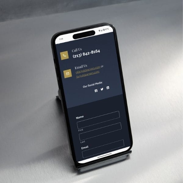 Smartphone displaying a contact page with a phone number, email addresses, and social media icons on a dark screen. The phone is on a stand placed on a gray surface.