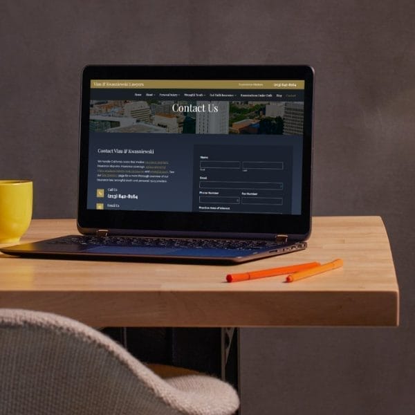 A laptop displaying a "Contact Us" webpage sits on a wooden table. A yellow mug and two orange pencils are next to the laptop. The background is a plain, gray wall.