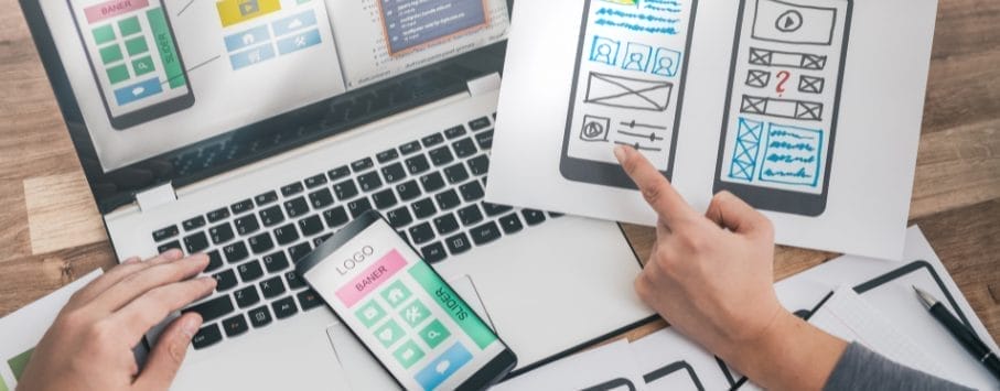A laptop on a desk displays design software. A person is holding a smartphone with a similar design. Another person points to a paper with a mobile app wireframe. Papers with sketches and designs are scattered around.