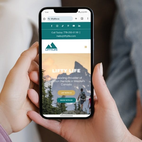 A person holding a smartphone showing a website for "Lifty Life," a vacation rental provider in Western Canada. The screen displays options to "List with Us" and "Book with Us," with mountain scenery in the background.