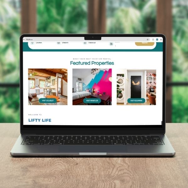 A laptop on a wooden table displays a vacation rental website with the title "Featured Properties" and images of various rental options, including a cozy cabin and a colorful room. Blurred greenery is visible through a window in the background.