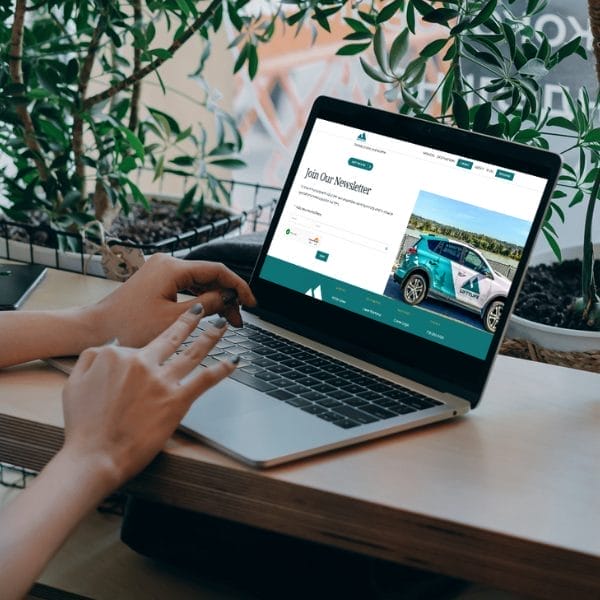 Person using a laptop at a wooden table with plants in the background. The laptop screen shows a webpage with a "Join Our Newsletter" form and an image of a car. The person is typing on the keyboard.