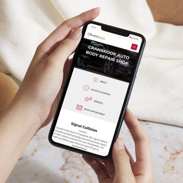 A person holding a smartphone displaying the Cranbrook Auto Body Repair Shop website. The screen shows navigation options like About, Hours & Location, Services, and Book Appointment. The background is a light fabric surface.
