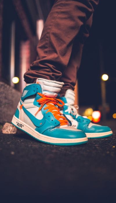 Close-up of a person wearing vibrant teal and white high-top sneakers with orange laces, standing on a city street at night. The background is blurred with streetlights and a slightly visible building. The person is wearing brown pants.