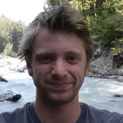 A person with light hair stands in front of a scenic river with a forested area in the background, smiling at the camera.