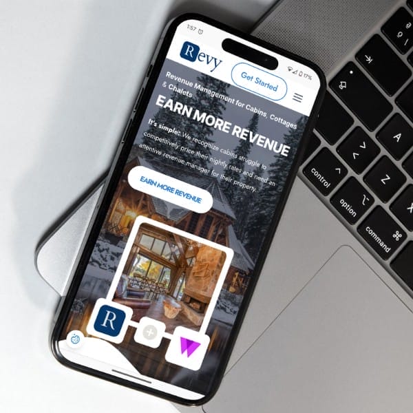Smartphone displaying a website for Revy, a revenue management service for cabins and cottages, is resting on a laptop. The screen shows an image of a snowy cabin with the text "Earn More Revenue" and buttons for getting started.