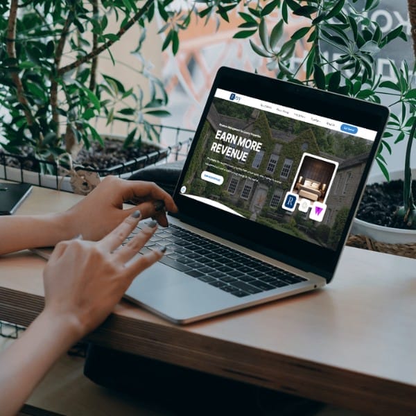 Person using a laptop displaying a website with the headline "Earn More Revenue." The laptop is on a wooden table surrounded by potted plants. Hands are typing on the keyboard.