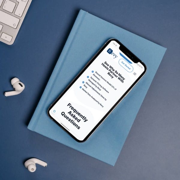 Smartphone displaying a webpage with frequently asked questions on a blue hardcover book, next to wireless earbuds and a computer keyboard on a blue surface.