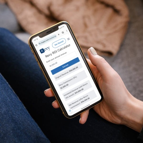 A person is holding a smartphone displaying a "Revy ROI Calculator" webpage. The screen shows a breakdown of revenues, commission, and net earnings. The person is seated on a sofa, revealing part of a hand and legs dressed in blue denim.