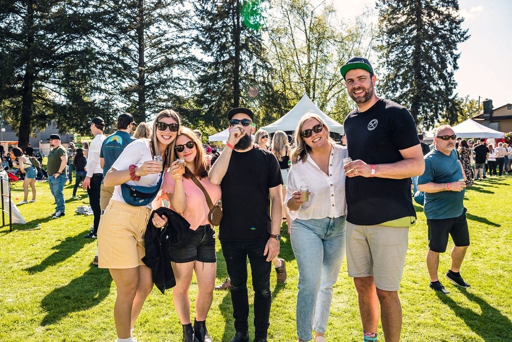 A group of five people posing and smiling outdoors at a sunny event. Tents and other attendees are visible in the background, with tall trees providing shade. Everyone is casually dressed and appears to be enjoying the day.