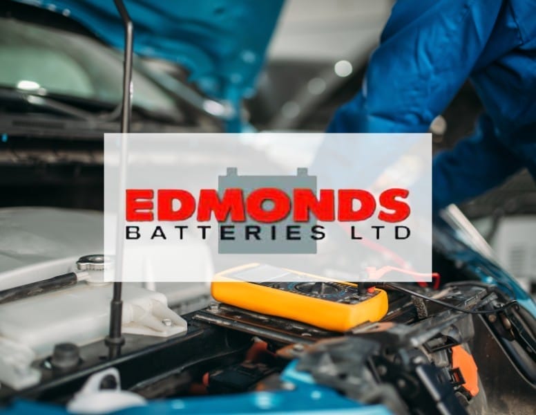 A mechanic in blue gloves uses a tool to check a car battery under the open hood of a vehicle. The image has a semi-transparent overlay with the text "EDMONDS BATTERIES LTD" in red and black letters.