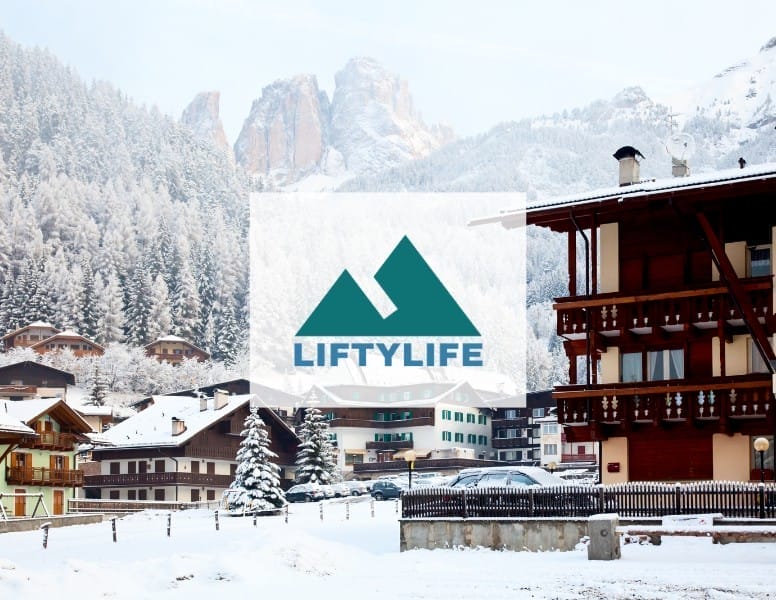 Snow-covered alpine village with wooden chalets, surrounded by pine trees and mountains in the background. A logo with two overlapping triangles and "LIFTYLIFE" is superimposed over the scene.