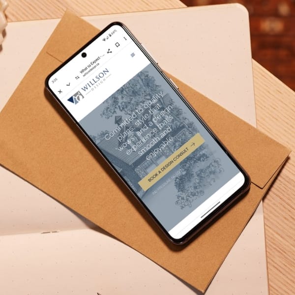 A smartphone displaying the "Willson" website rests on two brown envelopes atop a white surface. The screen shows text about design, alongside a button labeled "Book a Design Consult.