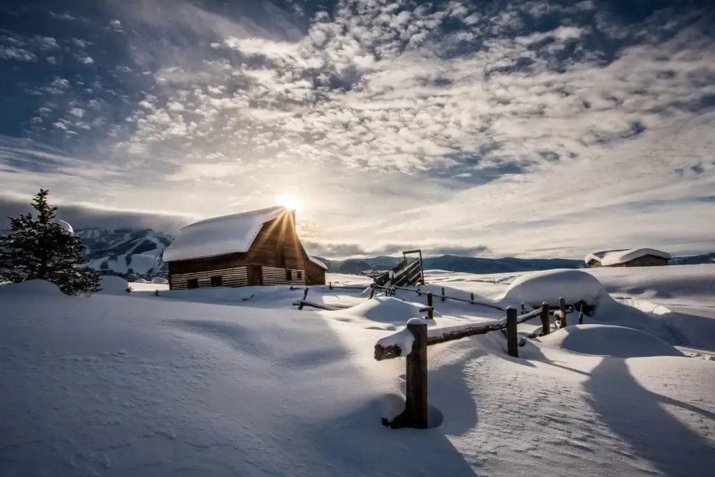 A snowy landscape features a rustic wooden cabin surrounded by deep snowdrifts under a sky dotted with clouds. The sun peeks behind the cabin, casting long shadows. In the distance, snow-covered mountains are visible.