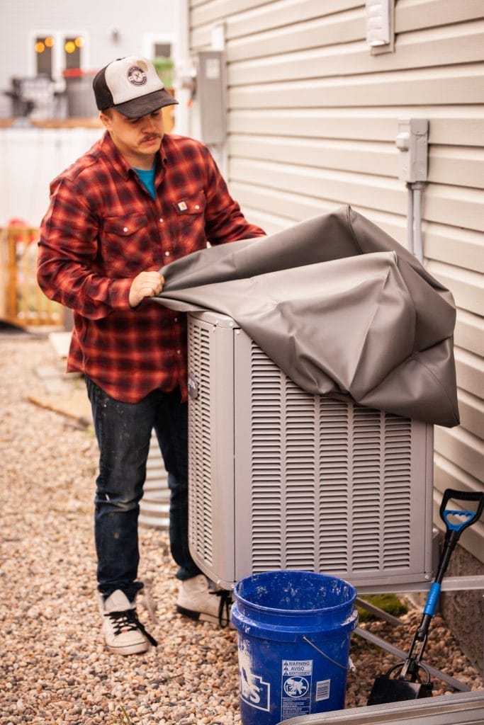 Person in a red plaid shirt and cap places a gray cover on an outdoor air conditioning unit beside a house. A blue bucket and shovel are on the gravel ground nearby.