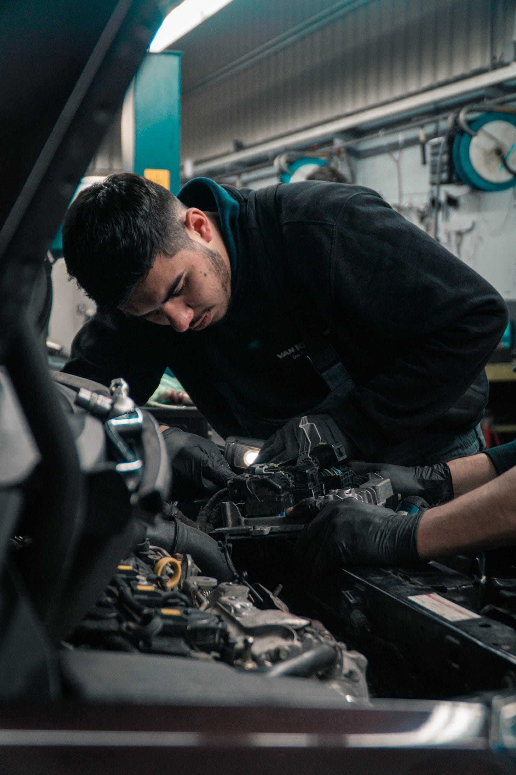 A person wearing a dark outfit works on a car engine in a garage, using a flashlight for visibility. The scene includes various tools and equipment in the background, highlighting a focused environment.