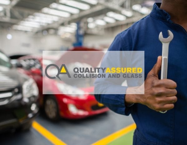 Mechanic in a blue jumpsuit holding a wrench, standing in an auto repair shop with cars in the background. Logo overlaid reads "Quality Assured Collision and Glass" in bold and italic font.