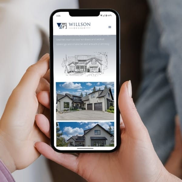 A person holding a smartphone displaying the Willson Design website. The screen shows images of modern house designs with gabled roofs. The website header contains the Willson Design logo and a navigation menu on an off-white background.