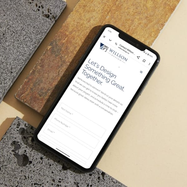 Smartphone displaying a website with a header "Let's Design Something Great. Together." rests on various stone tiles. The screen shows a contact form.