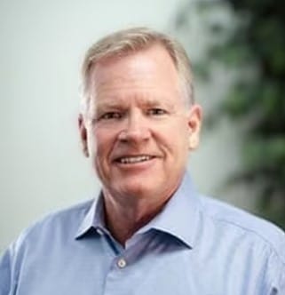 A middle-aged man with short, light hair wearing a light blue button-up shirt. He is smiling slightly, standing in front of a light background with some greenery visible.