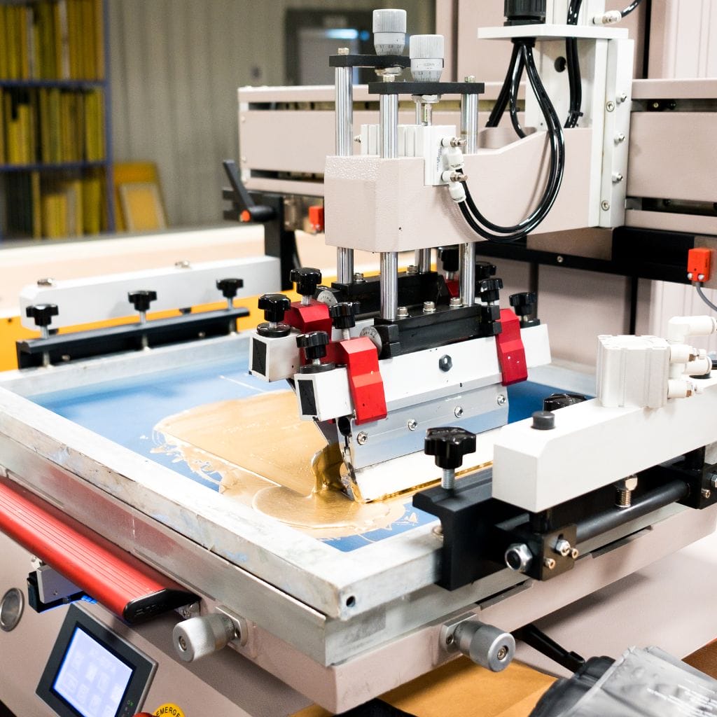 A close-up of a printing machine in operation, depositing brown ink onto a flat surface. The machine features various metal and plastic components, with shelves of yellow materials visible in the blurred background.