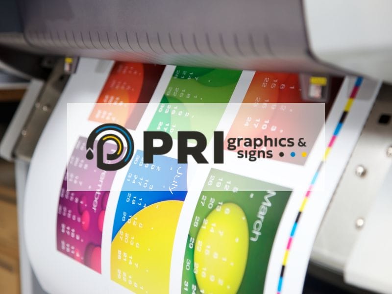 A large format printer outputs a color print showing a calendar with months of the year and colorful design elements. The image includes a semi-transparent overlay with the logo and text: "PRI graphics & signs.