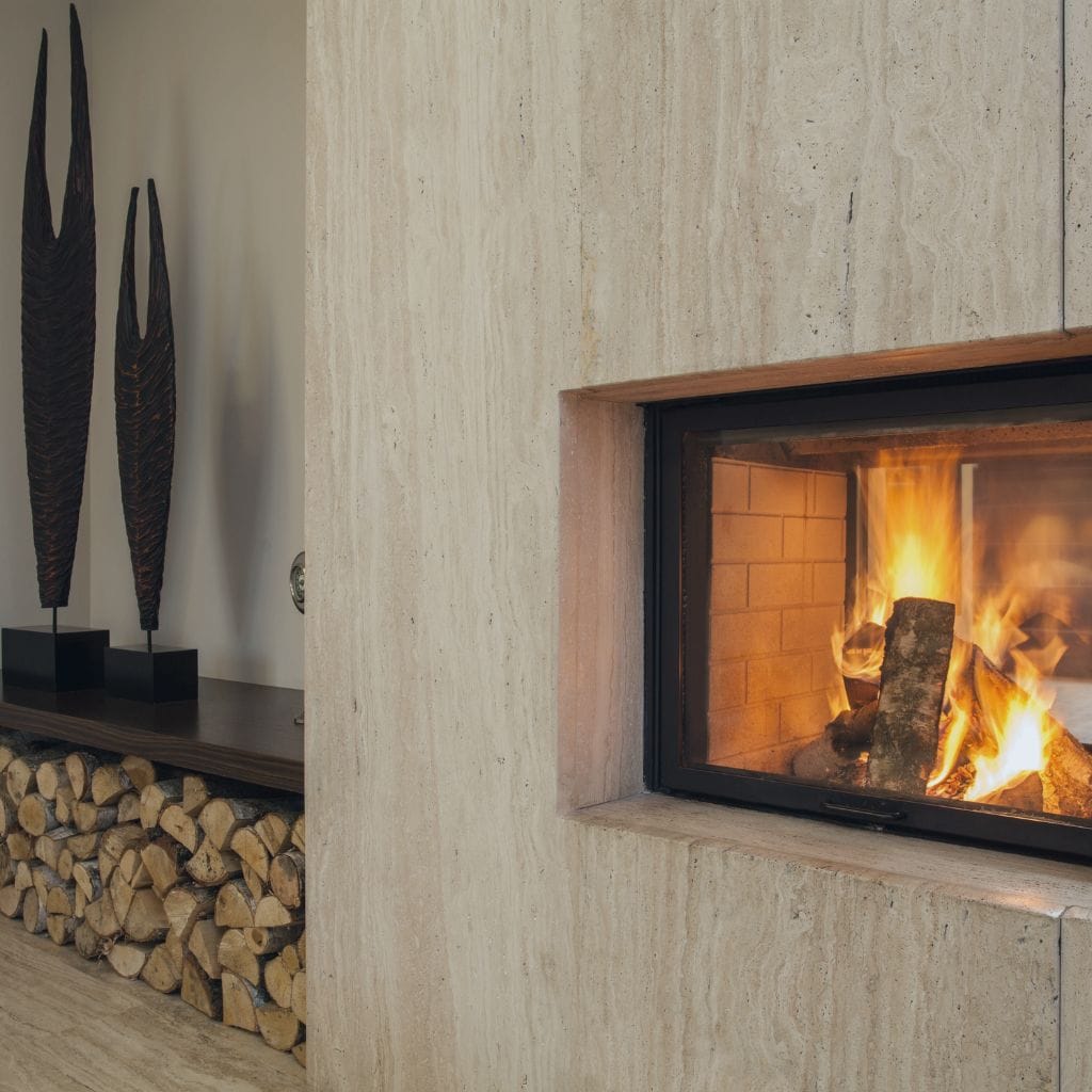 Modern living room with a lit fireplace set in a stone wall. Firewood is neatly stacked beneath the mantel. Two tall, abstract sculptures are displayed on a side table, adding a contemporary touch.