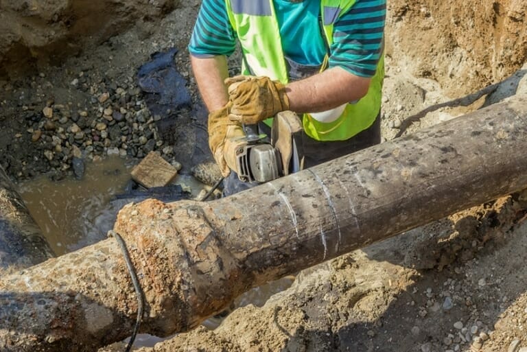 A construction worker in a blue and green striped shirt and safety vest uses a power tool to cut a large, rusty metal pipe in a muddy excavation site. He is wearing protective gloves as he works amid scattered gravel.