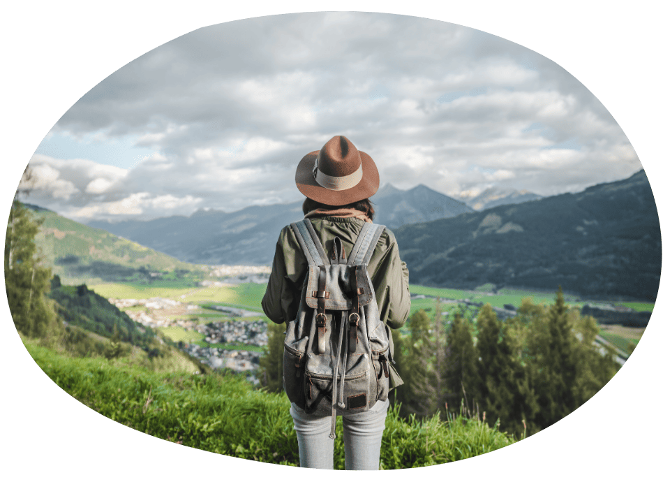 A person wearing a hat and backpack stands on a grassy hill, overlooking a scenic valley with mountains, greenery, and a cloudy sky in the background.