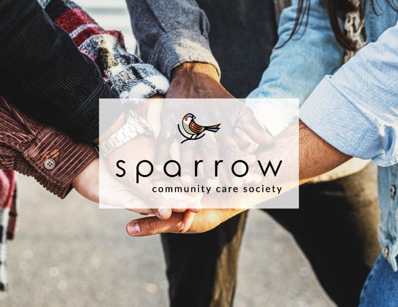 Four people join hands in unity. Overlaid is the Sparrow Community Care Society logo, featuring a sparrow illustration and the text "sparrow community care society." The background is slightly blurred outdoor scenery.