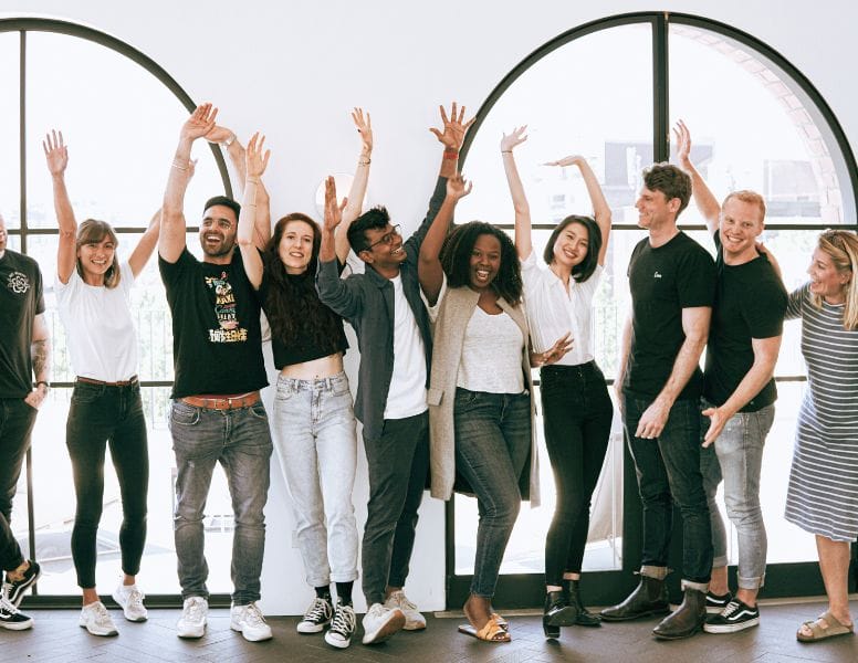 A diverse group of people stands in a line, smiling and raising their hands in a celebratory pose. They are standing indoors in front of large arched windows. Everyone is casually dressed, creating a joyful and relaxed atmosphere.