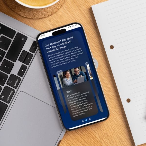 A smartphone on a wooden desk displays a webpage titled "Our Diamond Standard: Your Key to a Brilliant Benefit Strategy." It features an image of two people working on a laptop. Nearby are a silver laptop, an open notebook, and a coffee cup.