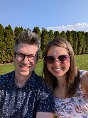 A man and a woman smiling outdoors. The man wears glasses and a patterned shirt, while the woman wears sunglasses and a floral dress. They are in front of a row of green trees under a clear blue sky.