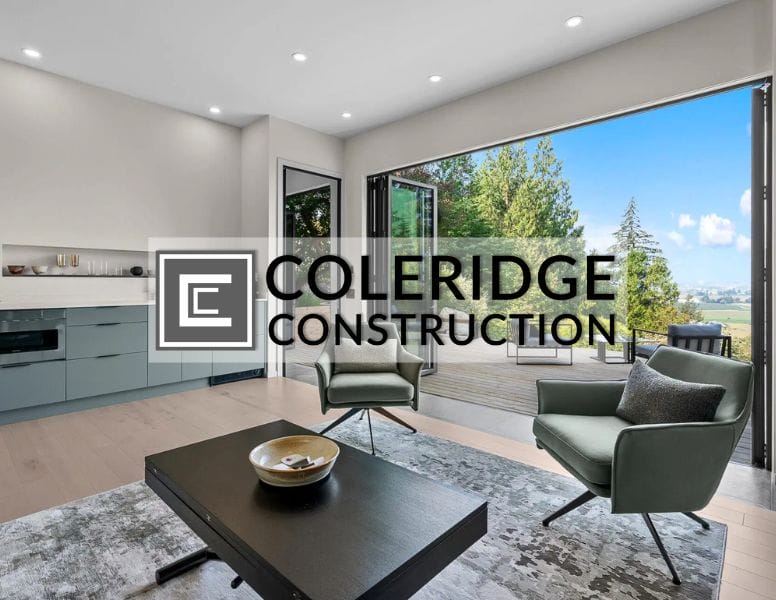A modern living room with green chairs, a large black coffee table, and a rug. The room opens to a spacious deck with a scenic view of trees and the sky. The text "Coleridge Construction" is overlaid on the image.