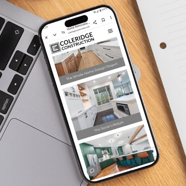 A smartphone displaying a construction company's website is resting on a silver laptop. The screen shows images of modern home interiors with text: "The Whole Home Design Project" and "The Social Lifestyle." A notepad and pen are nearby.