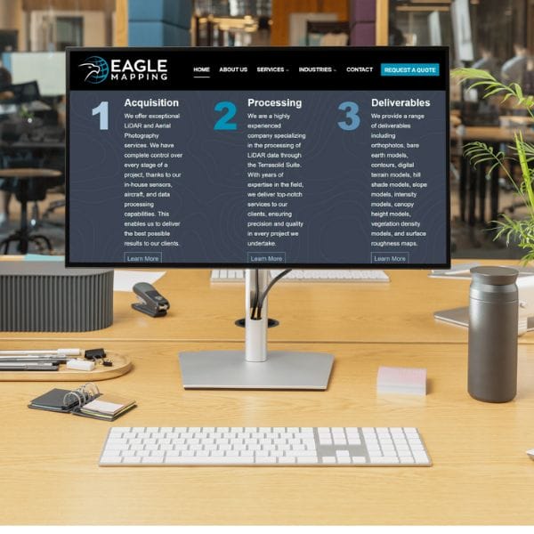 A computer monitor on a desk displays a website for "Eagle Mapping." The screen shows three columns titled "Acquisition," "Processing," and "Deliverables." The office setup includes a keyboard, tablet, and a thermal bottle.